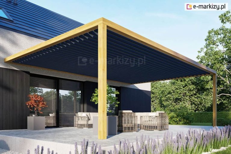 Zdjęcie prezentuje wizualizację nowoczesnego zadaszenia tarasu opartego na systemie lamelowym Selt SB400 MD. Konstrukcja nośna składa się z grubych, jasnych belek drewnianych, tworzących ramę wspartą na solidnym słupie narożnym. Wewnątrz ramy zamontowane są ruchome, aluminiowe lamele w kolorze ciemnego antracytu, które są ustawione pod lekkim kątem, co pozwala na regulację dostępu światła słonecznego. Pergola jest zintegrowana z bryłą nowoczesnego domu o ciemnej elewacji i dużych przeszkleniach. Pod zadaszeniem znajduje się strefa wypoczynkowa z jasnymi meblami ogrodowymi, a taras wyłożony jest szarymi płytami. Wokół widoczna jest zadbana roślinność, w tym lawenda na pierwszym planie oraz gęste drzewa w tle, co podkreśla luksusowy i wypoczynkowy charakter przestrzeni.