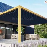 Drewniana pergola przyścienna z dachem lamelowym SB400 zamocowanym na jej konstrukcji osłania taras przy domu, którego elewacja została wykończona drewnem i cegłą. Pod pergolą ustawiono zestaw mebli wypoczynkowych w odcieniach brązu oraz dwie kwadratowe duże donice z małymi drzewkami: po lewej z czerwonymi, a pośrodku z zielonymi liśćmi. Przed tarasem wyłożonym płytkami gresowymi widoczny kwietnik z lawendą, a w tle widoczny żywopłot i wysokie drzewa liściaste oraz jasnoniebieskie niebo.