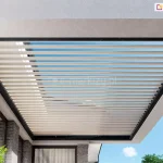 Widok z dołu na nowoczesne zadaszenie tarasu w formie dachu lamelowego Essential zintegrowanego z bryłą budynku. Konstrukcja zadaszenia składa się z solidnej, ciemnej ramy o grafitowym lub czarnym kolorze, w której osadzone są ruchome, poziome lamele w kolorze kremowym lub jasnobeżowym. Lamele są ustawione równolegle do siebie z zachowaniem niewielkich odstępów, przez które prześwituje jasnoniebieskie, bezchmurne niebo. Zadaszenie jest zamontowane pod betonowym, jasnym stropem, który stanowi przedłużenie dachu lub balkonu wyższej kondygnacji. Po lewej stronie widoczna jest elewacja domu wykonana z ciemnoszarej cegły klinkierowej oraz duże przeszklenia w ciemnych ramkach, w których odbija się otoczenie. W lewym dolnym rogu kadru znajdują się fragmenty jasnozielonej roślinności, prawdopodobnie liście juki lub ozdobnej trawy, które wprowadzają naturalny akcent do surowej, nowoczesnej architektury. Całość kompozycji podkreśla geometryczny porządek i minimalistyczny design systemu osłonowego.