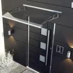 Minimalistyczne i nowoczesne zadaszenie wejścia wykonane z modelu Elegant, zamontowane na ciemnej, antracytowej elewacji budynku. Daszek posiada całkowicie transparentną, płaską płytę akrylową, która jest osadzona w subtelnych, srebrnych profilach stalowych mocowanych bezpośrednio do ściany. Konstrukcja osłania designerskie drzwi wejściowe w kolorze czarnym z poziomymi przetłoczeniami i mlecznymi przeszkleniami. Elewacja budynku wykonana jest z wielkoformatowych płyt o matowym wykończeniu, a nowoczesny charakter strefy wejściowej podkreślają symetrycznie rozmieszczone kinkiety LED rzucające ciepłe światło w górę i w dół oraz stalowa tabliczka z numerem posesji „1”. U podnóża ściany widoczny jest pas dekoracyjnego, jasnego żwiru oraz białe rośliny ozdobne (mrozy lub lawenda), co tworzy elegancki kontrast z ciemną architekturą obiektu.