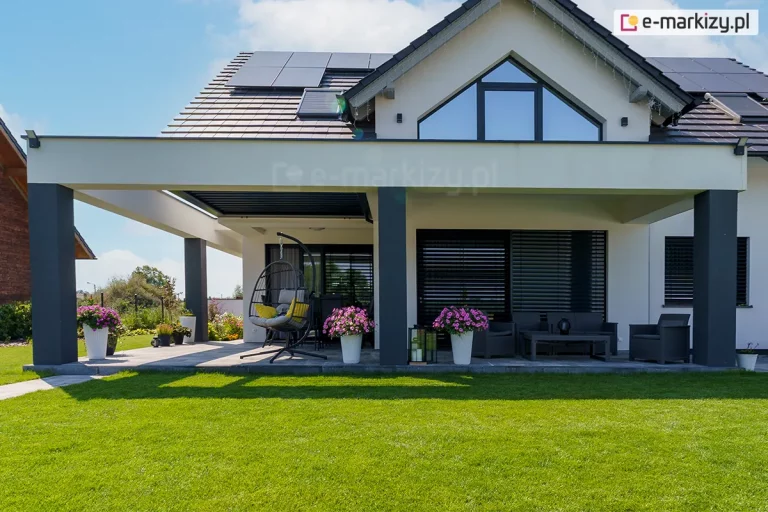 nowoczesny budynek jednorodzinny o jasnej elewacji i ciemnym, dwuspadowym dachu z zainstalowanymi panelami fotowoltaicznymi. Do bryły budynku przylega przestronny, zadaszony taras, którego konstrukcja opiera się na masywnych, ciemnoszarych słupach nośnych i białych belkach poprzecznych. Centralnym elementem zadaszenia jest system dachu lamelowego SB400R w kolorze antracytowym, który na fotografii jest całkowicie zamknięty, tworząc jednolitą płaszczyznę. Pod zadaszeniem zaaranżowano strefę wypoczynkową z szarymi meblami ogrodowymi, niskim stolikiem kawowym oraz wiszącym fotelem typu kokon. Taras wyłożony jest jasnoszarymi płytami betonowymi, na których ustawiono białe donice z obficie kwitnącymi, różowymi kwiatami. Przed tarasem rozpościera się zadbany, intensywnie zielony trawnik, a w oknach budynku widoczne są systemowe żaluzje fasadowe w kolorze antracytowym. Całość ujęcia charakteryzuje się czystą, minimalistyczną estetyką i wysokim standardem wykończenia.