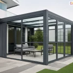 Nowoczesna realizacja ogrodu zimowego lub całorocznej zabudowy tarasu, zintegrowaną z bryłą minimalistycznego, białego budynku jednorodzinnego. Konstrukcja nośna została wykonana z grubych profili aluminiowych w kolorze antracytowym, które tworzą geometryczną ramę zadaszenia oraz ścian bocznych. W systemie wykorzystano szklane ściany stałe Fest, które dzielą przestrzeń na regularne sekcje za pomocą pionowych i poziomych szprosów, zapewniając pełną przezierność i ochronę przed warunkami atmosferycznymi. Dach konstrukcji jest w całości przeszklony, co pozwala na maksymalne doświetlenie wnętrza światłem dziennym. Wewnątrz przeszklonej strefy zaaranżowano miejsce do wypoczynku, wyposażone w komplet mebli tarasowych: dwuosobową sofę oraz fotel z szarymi, miękkimi poduchami, a także niski, prostokątny stolik kawowy z ciemnym blatem. Podłoże wewnątrz i wokół zabudowy stanowią wielkoformatowe płyty tarasowe w dwóch odcieniach szarości, ułożone w pasy. Po lewej stronie, przy ścianie budynku, widoczna jest pionowa przesłona materiałowa w formie bocznej rolety lub zasłony, zapewniająca dodatkową prywatność. Całość kompozycji dopełnia zadbany trawnik oraz niska roślinność ogrodowa widoczna w tle, co podkreśla spójność nowoczesnej architektury z otaczającą naturą.
