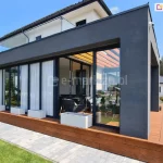 Zdjęcie przedstawia nowoczesną bryłę ocieplonego ogrodu zimowego o minimalistycznej architekturze, dobudowaną do piętrowego budynku mieszkalnego. Główną konstrukcję stanowią masywne ściany i zadaszenie wykończone ciemnoszarym tynkiem strukturalnym w kolorze antracytowym. Frontowa elewacja zabudowy jest niemal w całości przeszklona przy użyciu systemu przesuwnych drzwi ramowych Sotro w kolorze czarnym lub bardzo ciemnym graficie. Część skrzydeł jest przysłonięta od wewnątrz długimi, białymi firanami, co nadaje wnętrzu przytulny charakter. Całość konstrukcji posadowiona jest na podwyższonym fundamencie obłożonym drewnianą oblicówką w ciepłym, miodowym odcieniu, która tworzy niski cokół. Przed przeszkleniami ustawiono trzy białe, prostokątne donice o geometrycznych kształtach. Z boku zabudowy widoczne jest wejście na taras wyłożony deskami kompozytowymi w tym samym kolorze co cokół, prowadzące do wnętrza wyposażonego w ciemne meble wypoczynkowe. Nad częścią wypoczynkową, pod stałym dachem, zamontowano drewniane panele dekoracyjne lub lamele w kolorze drewna. Otoczenie budynku obejmuje starannie przystrzyżony trawnik, ścieżkę z szarej kostki brukowej oraz pas zieleni z regularnie rozmieszczonymi, formowanymi krzewami w kształcie kuli. W tle widoczny jest sąsiedni dom oraz ściana lasu pod bezchmurnym, błękitnym niebem.