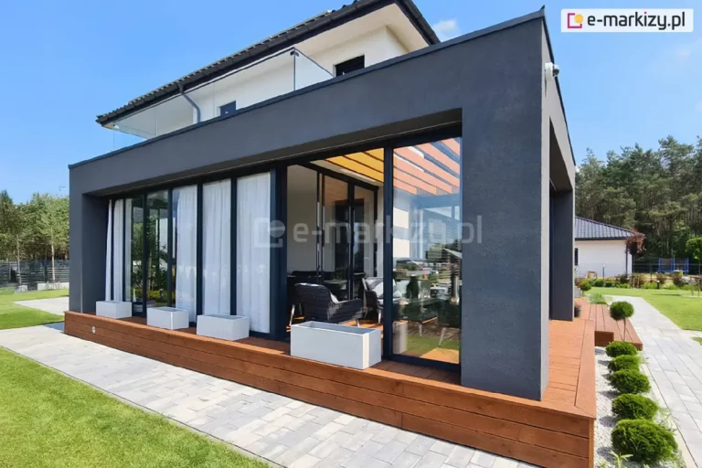 Zdjęcie przedstawia nowoczesną bryłę ocieplonego ogrodu zimowego o minimalistycznej architekturze, dobudowaną do piętrowego budynku mieszkalnego. Główną konstrukcję stanowią masywne ściany i zadaszenie wykończone ciemnoszarym tynkiem strukturalnym w kolorze antracytowym. Frontowa elewacja zabudowy jest niemal w całości przeszklona przy użyciu systemu przesuwnych drzwi ramowych Sotro w kolorze czarnym lub bardzo ciemnym graficie. Część skrzydeł jest przysłonięta od wewnątrz długimi, białymi firanami, co nadaje wnętrzu przytulny charakter. Całość konstrukcji posadowiona jest na podwyższonym fundamencie obłożonym drewnianą oblicówką w ciepłym, miodowym odcieniu, która tworzy niski cokół. Przed przeszkleniami ustawiono trzy białe, prostokątne donice o geometrycznych kształtach. Z boku zabudowy widoczne jest wejście na taras wyłożony deskami kompozytowymi w tym samym kolorze co cokół, prowadzące do wnętrza wyposażonego w ciemne meble wypoczynkowe. Nad częścią wypoczynkową, pod stałym dachem, zamontowano drewniane panele dekoracyjne lub lamele w kolorze drewna. Otoczenie budynku obejmuje starannie przystrzyżony trawnik, ścieżkę z szarej kostki brukowej oraz pas zieleni z regularnie rozmieszczonymi, formowanymi krzewami w kształcie kuli. W tle widoczny jest sąsiedni dom oraz ściana lasu pod bezchmurnym, błękitnym niebem.