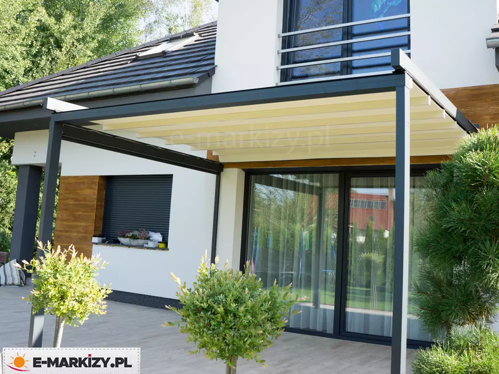 Na zdjęciu widoczna jest pergola tarasowa przyścienna Luxo, zamontowaną na elewacji piętrowego budynku o białych ścianach z drewnianymi akcentami. Konstrukcja pergoli wykonana jest z profili aluminiowych w kolorze antracytowym, wsparta na dwóch smukłych słupach frontowych. Zadaszenie stanowi ruchome poszycie tkaninowe w kolorze kremowym, które jest całkowicie rozwinięte i rozpięte na poprzecznych belkach przeciwwietrznych, tworząc płaską powierzchnię chroniącą przed słońcem. Pergola osłania duży, przeszklony otwór drzwiowy prowadzący na taras wyłożony jasnoszarymi płytami. Wokół tarasu widoczna jest starannie utrzymana roślinność, w tym formowane krzewy w donicach na pierwszym planie oraz wysokie iglaki po prawej stronie. Na elewacji budynku widoczne są również ciemne rolety zewnętrzne oraz barierka balkonowa na piętrze. Całość kompozycji podkreśla estetykę nowoczesnego budownictwa i funkcjonalność systemów przeciwsłonecznych w aranżacji przestrzeni wypoczynkowej.