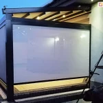 Zdjęcie prezentuje nowoczesną, przyścienną pergolę tarasową Square w warunkach wieczornych, z aktywnym systemem oświetlenia zintegrowanego. Konstrukcja pergoli wykonana jest z profili aluminiowych w kolorze antracytowym i posiada jednospadowy dach. Pod poszyciem tkaninowego dachu zamontowano poprzeczne belki, w których umieszczono punktowe źródła światła LED, emitujące ciepły blask na przestrzeń tarasu. Frontowa oraz boczna ściana pergoli zostały osłonięte pionowymi roletami typu screen w kolorze białym lub bardzo jasnoszarym; rolety są całkowicie opuszczone, tworząc półtransparentną barierę wizualną. Wnętrze pergoli jest rozświetlone, co pozwala dostrzec zarys jasnej posadzki wykonanej z kostki brukowej. Po prawej stronie, na zewnątrz konstrukcji, znajduje się metalowe palenisko ogrodowe lub trójnóg z rusztem. Pergola przylega do jasnej elewacji budynku z widocznym oknem, w którym odbija się wewnętrzne oświetlenie domu. Całość ujęcia charakteryzuje się wysokim kontrastem między ciemną bryłą konstrukcji a rozświetlonymi płaszczyznami materiału osłonowego na tle zapadającego zmroku.
