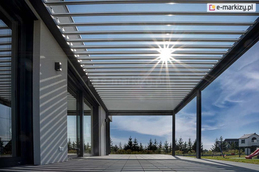 Promienie słoneczne prześwitujące przez uchylone pióra lameli dużej pergoli ogrodowej na tle niebieskiego nieba z chmurkami