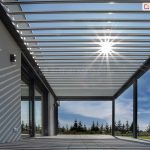 Promienie słoneczne prześwitujące przez uchylone pióra lameli dużej pergoli ogrodowej na tle niebieskiego nieba z chmurkami