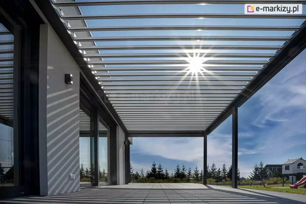 Duża pergola lamelowa przyścienna SB400 Aluprof z antracytową konstrukcją i białymi, uchylonymi lamelami nad dużym tarasem przy nowoczesnym budynku wyłożonym płytkami w jasnoszarym kolorze. W tle widoczne drzewka iglaste oraz sąsiedni dom pod niebieskim niebem z postrzępionymi chmurami. Przez uchylone lamele pergoli wpada światło słoneczne.