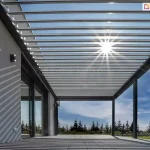 Duża pergola lamelowa przyścienna SB400 Aluprof z antracytową konstrukcją i białymi, uchylonymi lamelami nad dużym tarasem przy nowoczesnym budynku wyłożonym płytkami w jasnoszarym kolorze. W tle widoczne drzewka iglaste oraz sąsiedni dom pod niebieskim niebem z postrzępionymi chmurami. Przez uchylone lamele pergoli wpada światło słoneczne.