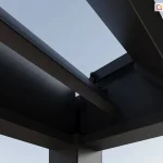 Zbliżenie na górny narożnik konstrukcji antracytowej pergoli lamelowej Marlo, ilustrujące mechanizm obrotowy lameli. Widoczne są dwa główne profile ramy łączące się pod kątem prostym, osadzone na pionowym słupie o kwadratowym przekroju. Wewnątrz ramy zamontowane są ruchome lamele o aerodynamicznym kształcie, które na zdjęciu znajdują się w pozycji otwartej, odsłaniając jasne niebo. Na krawędzi profilu bocznego widoczne są punkty mocowania oraz elementy mechaniczne odpowiedzialne za płynną zmianę kąta nachylenia piór. Konstrukcja charakteryzuje się matowym wykończeniem powierzchni oraz precyzyjnym spasowaniem elementów aluminiowych. Detal ten podkreśla techniczną stronę systemu bioklimatycznego, umożliwiającego regulację dopływu światła i cyrkulację powietrza pod zadaszeniem.