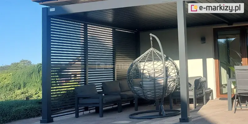 Zdjęcie prezentuje szerokie ujęcie nowoczesnej pergoli tarasowej w kolorze antracytowym, zainstalowanej przy ścianie budynku. Konstrukcja posiada płaski dach z poziomymi lamelami, a jej lewy bok został w całości zabudowany trzema pionowymi panelami żaluzjowymi. Panele te składają się z gęsto rozmieszczonych poziomych listew; dwa skrajne panele mają lamele ustawione pod kątem, co tworzy prześwity i pozwala na wgląd do ogrodu, natomiast ostatni, umieszczony najbliżej ściany panel wydaje się być bardziej domknięty. Pod zadaszeniem zaaranżowano strefę wypoczynkową z meblami ogrodowymi. Widoczna jest niska, ciemna sofa modułowa z grubymi, grafitowymi poduszkami, ustawiona wzdłuż żaluzjowej ściany. Na pierwszym planie, nieco po prawej stronie od środka, znajduje się wiszący fotel typu kokon w kolorze ciemnoszarym, zawieszony na wygiętym pałąku stelaża. Wewnątrz fotela ułożone są jasnoszare poduszki. Po prawej stronie kadru widoczny jest fragment elewacji domu w jasnym, kremowym kolorze z dużym przeszkleniem drzwi tarasowych oraz metalowa konstrukcja stołu z krzesłem. Podłoże tarasu wyłożone jest estetyczną kostką brukową w odcieniach beżu i szarości. W tle, po lewej stronie za pergolą, rozciąga się widok na gęstą, zieloną roślinność ogrodową pod bezchmurnym, błękitnym niebem.