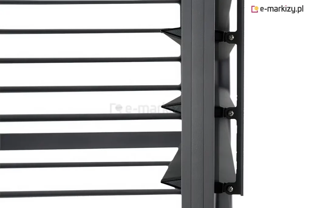 Zbliżenie na techniczny detal konstrukcyjny ruchomych lameli bocznego panelu żaluzjowego do pergoli tarasowej w kolorze antracytowym. Na zdjęciu widoczny jest pionowy profil ramy, do którego zamocowano mechanizm obrotowy poziomych listew. Lamele mają charakterystyczny, aerodynamiczny kształt przypominający spłaszczony romb lub klin, co zapewnia ich szczelne przyleganie po zamknięciu. Mocowanie każdej z listew odbywa się za pomocą czarnych, polimerowych uchwytów przykręconych do bocznej szyny za pomocą srebrnych wkrętów. Po prawej stronie profilu widoczna jest pionowa listwa sterująca, która łączy poszczególne uchwyty lameli, umożliwiając ich synchroniczny obrót i regulację kąta nachylenia. Powierzchnia aluminiowych elementów posiada matowe, lekko ziarniste wykończenie typu strukturalnego. Tło za konstrukcją jest jednolicie białe, co pozwala na wyraźne wyeksponowanie detali mechanicznych i precyzji wykonania systemu żaluzjowego.
