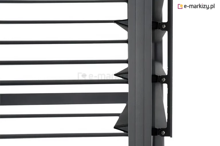 Zbliżenie na techniczny detal konstrukcyjny ruchomych lameli bocznego panelu żaluzjowego do pergoli tarasowej w kolorze antracytowym. Na zdjęciu widoczny jest pionowy profil ramy, do którego zamocowano mechanizm obrotowy poziomych listew. Lamele mają charakterystyczny, aerodynamiczny kształt przypominający spłaszczony romb lub klin, co zapewnia ich szczelne przyleganie po zamknięciu. Mocowanie każdej z listew odbywa się za pomocą czarnych, polimerowych uchwytów przykręconych do bocznej szyny za pomocą srebrnych wkrętów. Po prawej stronie profilu widoczna jest pionowa listwa sterująca, która łączy poszczególne uchwyty lameli, umożliwiając ich synchroniczny obrót i regulację kąta nachylenia. Powierzchnia aluminiowych elementów posiada matowe, lekko ziarniste wykończenie typu strukturalnego. Tło za konstrukcją jest jednolicie białe, co pozwala na wyraźne wyeksponowanie detali mechanicznych i precyzji wykonania systemu żaluzjowego.