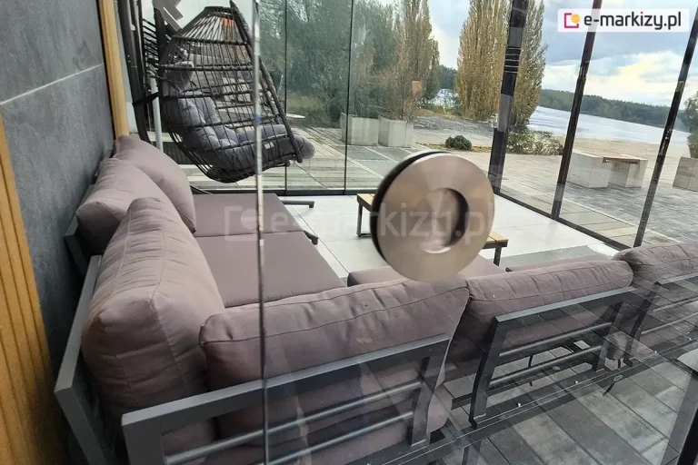 Zadaszenie tarasu wyposażone w system szklanych ścian przesuwnych dookoła. Na pierwszym planie widoczny srebrny metalowych uchwyt zamocowany w panelu auris. W tle pod zadaszeniem widoczne meble wypoczynkowe: niski narożnik z poduszkami oraz fotel typu kokon. Za ścianami zadaszenia widoczne betonowe patio oraz położona w głębi plaża nad jeziorem.