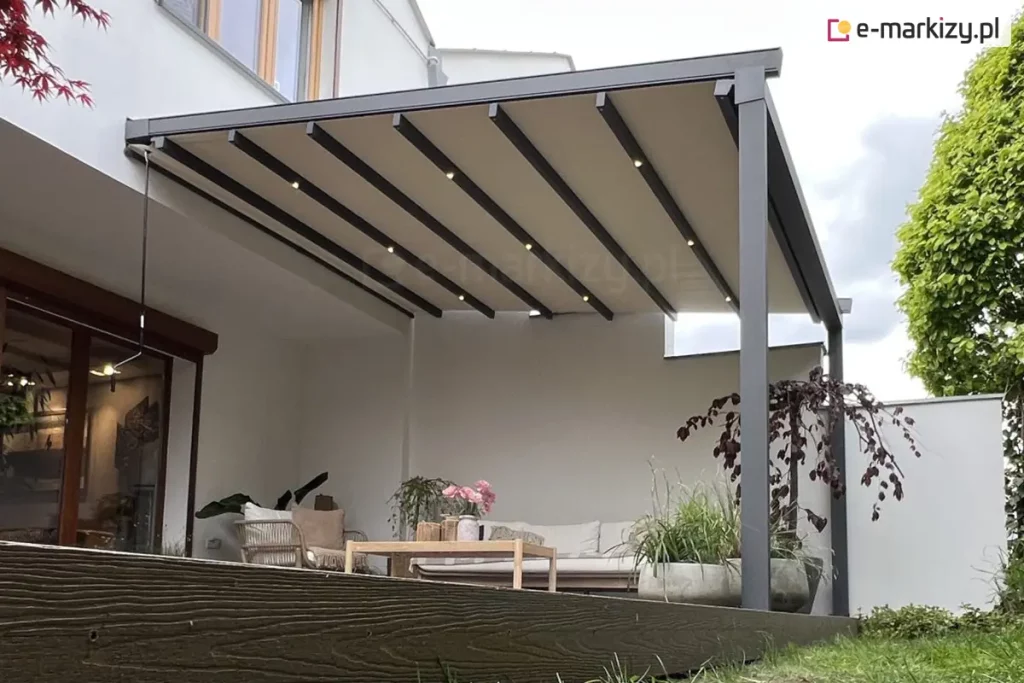 Ujęcie z perspektywy od dołu ukazuje Nowoczesną pergolę tarasową o antracytowej, aluminiowej konstrukcji przyściennej, zamontowaną do białej elewacji budynku. Zadaszenie składa się z beżowego materiału technicznego rozpiętego na poprzecznych profilach, w których zamontowano zintegrowane, punktowe oświetlenie LED emitujące ciepłe światło. Konstrukcja wsparta jest na solidnych, kwadratowych słupach bocznych. Taras, wykonany z ciemnobrązowych desek o wyraźnej strukturze drewna, znajduje się na lekkim podwyższeniu względem przylegającego do niego trawnika. W strefie wypoczynkowej pod pergolą umieszczono jasną sofę, fotel oraz niski drewniany stolik kawowy, na którym stoi wazon z różowymi kwiatami. Aranżację uzupełniają duże, szare donice o obłych kształtach, obsadzone zielonymi trawami ozdobnymi oraz drzewkiem o ciemnobordowych liściach. Po prawej stronie widoczny jest fragment wysokiego, gęstego żywopłotu, a w tle biały mur oddzielający posesję. Zdjęcie wykonane w dzień, przy lekko zachmurzonym niebie, co podkreśla nowoczesny i minimalistyczny charakter zabudowy.