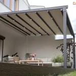 Ujęcie z perspektywy od dołu ukazuje Nowoczesną pergolę tarasową o antracytowej, aluminiowej konstrukcji przyściennej, zamontowaną do białej elewacji budynku. Zadaszenie składa się z beżowego materiału technicznego rozpiętego na poprzecznych profilach, w których zamontowano zintegrowane, punktowe oświetlenie LED emitujące ciepłe światło. Konstrukcja wsparta jest na solidnych, kwadratowych słupach bocznych. Taras, wykonany z ciemnobrązowych desek o wyraźnej strukturze drewna, znajduje się na lekkim podwyższeniu względem przylegającego do niego trawnika. W strefie wypoczynkowej pod pergolą umieszczono jasną sofę, fotel oraz niski drewniany stolik kawowy, na którym stoi wazon z różowymi kwiatami. Aranżację uzupełniają duże, szare donice o obłych kształtach, obsadzone zielonymi trawami ozdobnymi oraz drzewkiem o ciemnobordowych liściach. Po prawej stronie widoczny jest fragment wysokiego, gęstego żywopłotu, a w tle biały mur oddzielający posesję. Zdjęcie wykonane w dzień, przy lekko zachmurzonym niebie, co podkreśla nowoczesny i minimalistyczny charakter zabudowy.