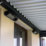 Zbliżenie na detale techniczne wyposażenia dodatkowego pergoli Essential Tarasola, zamontowanego pod jej zadaszeniem. Na jasnej, tynkowanej elewacji budynku widoczny jest nowoczesny, podłużny promiennik ciepła w czarnej obudowie, umieszczony bezpośrednio pod belką konstrukcyjną pergoli. Promiennik posiada metalową kratkę osłaniającą element grzejny i jest skierowany pod kątem w stronę strefy wypoczynkowej. W tle, wzdłuż tej samej ściany, zamontowane jest drugie, analogiczne urządzenie grzewcze. Powyżej widoczny jest fragment dachu lamelowego modelu Essential Tarasola wykonanego z białych, aluminiowych profili. Lamele są ustawione w pozycji zamkniętej, tworząc szczelną, ząbkowaną krawędź na styku z ciemnoantracytową ramą nośną. Konstrukcja ramy charakteryzuje się matowym wykończeniem i solidnym profilem. W dolnej części kadru widoczne jest brązowe obramowanie okna tarasowego, a w prawym dolnym rogu – fragment pionowego słupa i rynny odprowadzającej wodę. Zdjęcie podkreśla wysoką jakość montażu akcesoriów dedykowanych do systemu Essential Tarasola, które zwiększają komfort użytkowania tarasu w chłodniejsze dni.