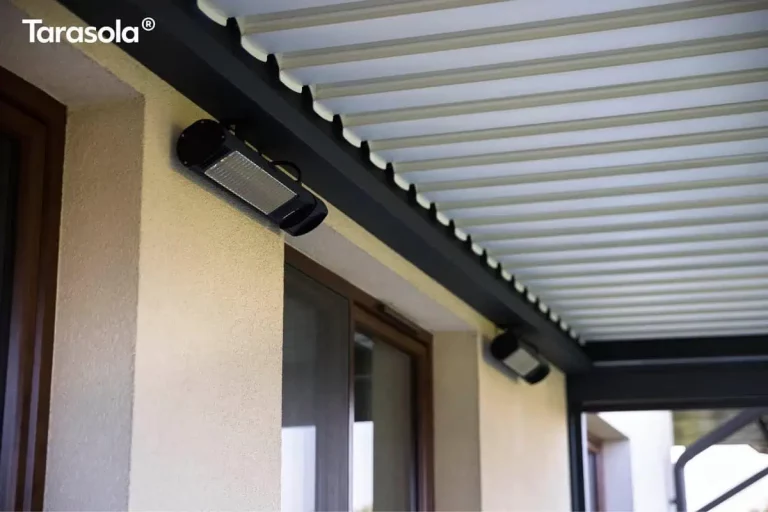 Zbliżenie na detale techniczne wyposażenia dodatkowego pergoli Essential Tarasola, zamontowanego pod jej zadaszeniem. Na jasnej, tynkowanej elewacji budynku widoczny jest nowoczesny, podłużny promiennik ciepła w czarnej obudowie, umieszczony bezpośrednio pod belką konstrukcyjną pergoli. Promiennik posiada metalową kratkę osłaniającą element grzejny i jest skierowany pod kątem w stronę strefy wypoczynkowej. W tle, wzdłuż tej samej ściany, zamontowane jest drugie, analogiczne urządzenie grzewcze. Powyżej widoczny jest fragment dachu lamelowego modelu Essential Tarasola wykonanego z białych, aluminiowych profili. Lamele są ustawione w pozycji zamkniętej, tworząc szczelną, ząbkowaną krawędź na styku z ciemnoantracytową ramą nośną. Konstrukcja ramy charakteryzuje się matowym wykończeniem i solidnym profilem. W dolnej części kadru widoczne jest brązowe obramowanie okna tarasowego, a w prawym dolnym rogu – fragment pionowego słupa i rynny odprowadzającej wodę. Zdjęcie podkreśla wysoką jakość montażu akcesoriów dedykowanych do systemu Essential Tarasola, które zwiększają komfort użytkowania tarasu w chłodniejsze dni.