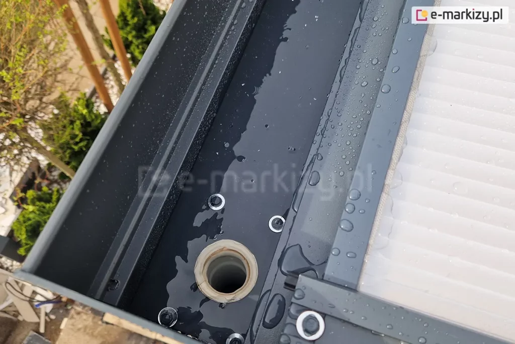 Zbliżenie połączenia rynny w belce przedniej zadaszenia Modes z rurą spustową ukrytą w nodze tego zadaszenia. Całość tworzy spójny system odprowadzania deszczówki z powierzchni poliwęglanu i zapewnia szczelną osłonę tarasu przed deszczem.