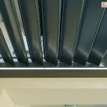 Dach lamelowy do pergoli w antracytowym kolorze model SB400 R Selt z zainstalowanym do ramy odpływem wody, ukrytym w ścianie. Uchylone czarne lamele kontrastują z listwą oświetlenia ledowego oraz białymi ścianami tarasu.