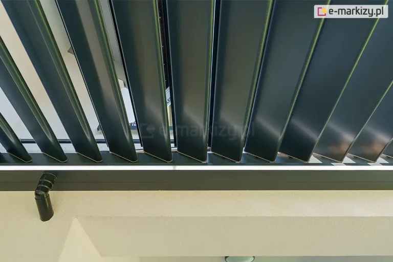 Dach lamelowy do pergoli w antracytowym kolorze model SB400 R Selt z zainstalowanym do ramy odpływem wody, ukrytym w ścianie. Uchylone czarne lamele kontrastują z listwą oświetlenia ledowego oraz białymi ścianami tarasu.