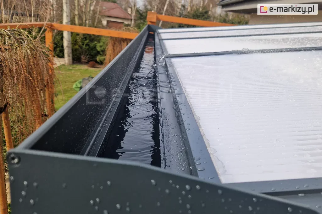 Zbliżenie rynny odpływowej zadaszenia z poliwęglanu Modes, ukrytej w belce przedniej, która wypełniona jest wodą. Woda z płyt poliwęglanowych zadaszenia spływa do przodu, skąd odprowadzana jest do podpór a nimi do ziemi lub na trawnik. W tle widoczny ogród z roślinami i dwiema drewnianymi konstrukcjami ozdobnymi.