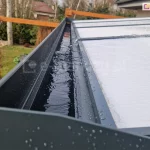 Zbliżenie rynny odpływowej zadaszenia z poliwęglanu Modes, ukrytej w belce przedniej, która wypełniona jest wodą. Woda z płyt poliwęglanowych zadaszenia spływa do przodu, skąd odprowadzana jest do podpór a nimi do ziemi lub na trawnik. W tle widoczny ogród z roślinami i dwiema drewnianymi konstrukcjami ozdobnymi.