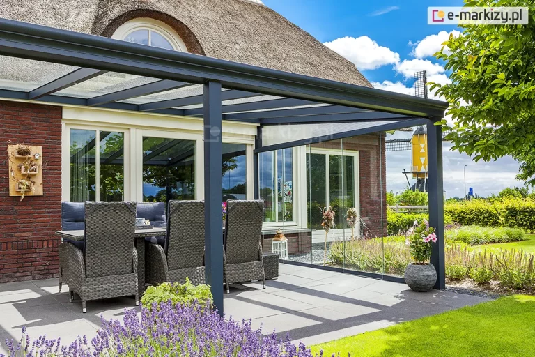 Nowoczesny ogród letni przylegający do budynku z ceglaną elewacją i dachem krytym strzechą. Zabudowa opiera się na aluminiowej konstrukcji zadaszenia Terano w kolorze antracytowym, która została uzupełniona o system bocznych szklanych drzwi przesuwnych. Transparentne panele dachu i ścian bocznych zapewniają pełny dostęp światła dziennego. Na tarasie wyłożonym wielkoformatowymi szarymi płytami znajduje się stół z wysokimi fotelami technorattanowymi wyposażonymi w niebieskie poduszki. Przed tarasem rośnie lawenda oraz inne rośliny ozdobne w donicach. W tle, za szklaną zabudową, widoczny jest wiejski krajobraz z tradycyjnym wiatrakiem typu koźlak pod błękitnym niebem z białymi chmurami.