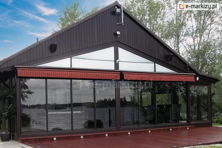 Zdjęcie przedstawia nowoczesną zabudowę tarasu restauracyjnego w formie ogrodu letniego, przylegającą do ciemnobrązowego budynku o konstrukcji szczytowej. Głównym elementem są wielkoformatowe, przesuwne przeszklenia ramowe typu Sotro, które tworzą transparentną ścianę frontową, odbijającą widok na pobliskie jezioro i linię drzew. Nad szklanymi panelami zamontowano dwie poziome markizy kasetowe z tkaniną w kolorze ceglastym lub ciemnoczerwonym w delikatne pasy, które pełnią funkcję osłony przeciwsłonecznej. Powyżej linii markiz, w szczytowej części elewacji, znajdują się trójkątne okna wyposażone w białe plisy dopasowane do kształtu ramy. Całość konstrukcji oparta jest na ciemnych profilach harmonizujących z drewnianą elewacją budynku. Przed ogrodem letnim rozciąga się przestronny taras wykonany z desek kompozytowych lub drewna w odcieniu mahoniowym, w który wbudowano punktowe oprawy oświetleniowe typu LED rozmieszczone wzdłuż krawędzi przy przeszkleniach. Po lewej stronie widoczny jest fragment dekoracyjnej roślinności w donicy, a w tle rozpościera się naturalny krajobraz z akwenem wodnym pod lekko zachmurzonym niebem.