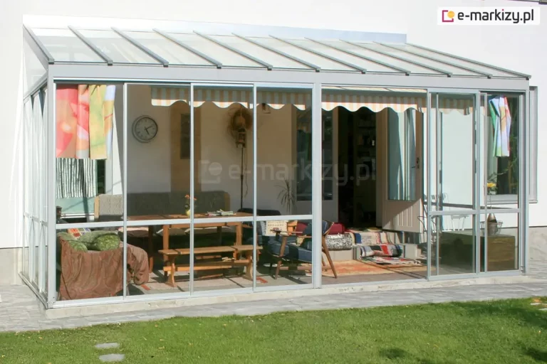 Zdjęcie przedstawia przyścienną konstrukcję ogrodu zimowego o lekkiej, aluminiowej budowie w kolorze jasnoszarym, zamontowaną na betonowym podwyższeniu. Zadaszenie obiektu jest jednospadowe, wykonane z przezroczystych paneli szklanych rozdzielonych profilami, pod którymi zainstalowano markizę tarasową w pasy o jasnej kolorystyce. Ściany frontowa i boczna są w pełni przeszklone i wyposażone w system drzwi przesuwnych osadzonych w ramach, co pozwala na swobodne otwieranie przestrzeni. Wewnątrz znajduje się strefa wypoczynkowa z drewnianym stołem, ławami oraz sofą przykrytą kocami, a na ścianie budynku widoczny jest okrągły zegar analogowy. Całość konstrukcji graniczy bezpośrednio z zadbanym trawnikiem, a wejście do wnętrza domu prowadzi przez białe, otwarte drzwi balkonowe.