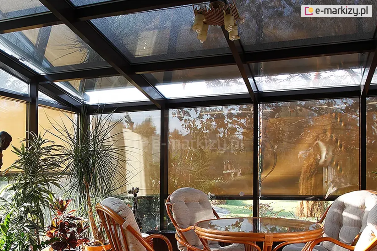 Wnętrze ogrodu zimowego o konstrukcji z antracytowych profili aluminiowych, wyposażone w system pionowych i dachowych osłon przeciwsłonecznych. Przeszklone ściany boczne osłonięte zostały refleksolami, a nad dachem zainstalowano system veranda, które rozpraszają światło słoneczne. W pomieszczeniu znajdują się meble rattanowe: okrągły stół ze szklanym blatem oraz fotele z jasnymi, pikowanymi poduszkami. Przestrzeń udekorowana jest licznymi roślinami doniczkowymi, w tym dużą draceną i krotonem. Z sufitu zwisa dekoracyjna lampa, a przez tkaninę rolet prześwituje ogród z drzewami i zielonym trawnikiem.