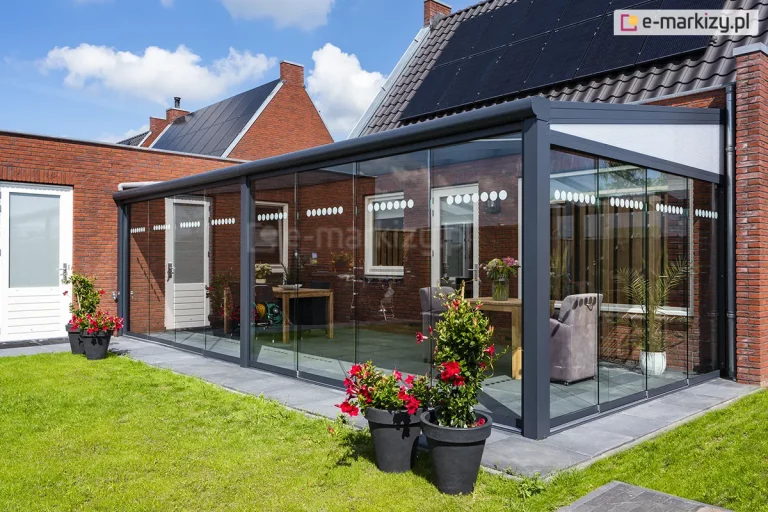 Zewnętrzny widok na nowoczesny ogród zimowy stworzony poprzez pełną zabudowę zadaszenia tarasowego Lunaro. Konstrukcja w kolorze antracytowym, z jednospadowym dachem wypełnionym panelami z poliwęglanu, jest wyposażona w system szklanych ścian przesuwnych Auris, które chronią wnętrze tarasu przed wiatrem i deszczem. Na szklanych taflach umieszczono dekoracyjne pasy białych kółek, pełniące funkcję estetyczną i ostrzegawczą. Wewnątrz widoczne są meble ogrodowe, w tym stół jadalniany i tapicerowane fotele, a przed konstrukcją, na tle zielonego trawnika, ustawiono duże czarne donice z kwitnącymi na czerwono roślinami. Budynek, do którego przylega konstrukcja, wykonany jest z czerwonej cegły i posiada dach pokryty czarną dachówką z zamontowanymi panelami fotowoltaicznymi.