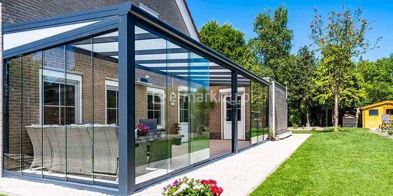 Boczne ujęcie całkowicie zabudowanego tarasu w formie ogrodu zimowego, przylegającego do ściany piętrowego budynku z jasnobrązowej cegły. Konstrukcja wykonana jest z profili aluminiowych w kolorze antracytowym, a zadaszenie pokrywają płyty z poliwęglanu komorowego w kolorze mlecznym. Ściany boczne oraz frontowe zabudowy stanowią przesuwne panele szklane Auris, które są całkowicie zamknięte, tworząc szczelną przestrzeń. Wewnątrz widoczna jest strefa wypoczynkowa z dużą, plecioną sofą narożną w kolorze jasnoszarym. Całość posadowiona jest na tarasie wyłożonym drobnym, jasnym kruszywem, który graniczy z pasem białych kamieni ozdobnych i równo przystrzyżonym trawnikiem. W tle widoczny jest ogród z gęstą roślinnością, drzewami oraz małym, żółtym domkiem narzędziowym w oddali. Na elewacji budynku widoczne są białe ramy okienne i drzwiowe z zewnętrznymi roletami, a na pierwszym planie znajduje się fragment donicy z kolorowymi kwiatami.