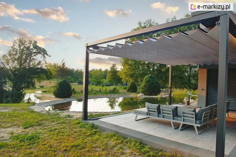 Szeroki kadr prezentujący nowoczesną pergolę tarasową Tako zamontowaną przy budynku, stanowiącą luksusową strefę wypoczynku nad brzegiem naturalnego stawu. Konstrukcja pergoli w kolorze antracytowym wyposażona jest w jasnobeżowe zadaszenie rzymskie, które jest częściowo rozsunięte, tworząc faliste linie. Pod zadaszeniem, na tarasie wyłożonym dużymi, jasnoszarymi płytami, znajduje się zestaw mebli ogrodowych – szary narożnik z miękkimi poduszkami. W tle widoczny jest malowniczy krajobraz z oczkiem wodnym lub stawem, w którego tafli odbijają się okoliczne drzewa i niebo. Brzeg stawu jest naturalnie uformowany, otoczony zieloną roślinnością, krzewami i drzewami liściastymi. Niebo w kolorze błękitnym z delikatnymi chmurami sugeruje porę późnego popołudnia (złota godzina), co nadaje scenie ciepłe, nastrojowe oświetlenie. Całość kompozycji podkreśla estetykę połączenia nowoczesnej architektury ogrodowej z naturalnym otoczeniem.