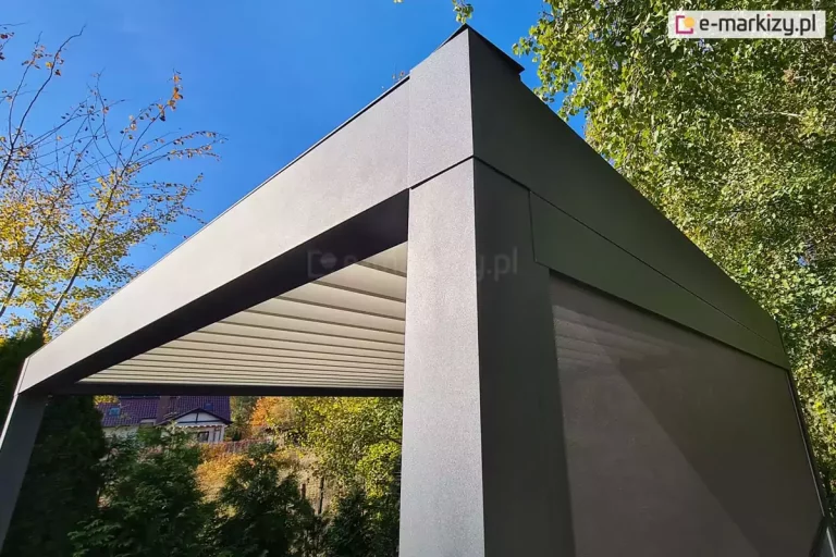 Fotografia prezentuje detal konstrukcyjny narożnika nowoczesnej pergoli tarasowej, wykonanej z grubych profili aluminiowych o matowej, ziarnistej strukturze w kolorze ciemnego grafitu. Na pierwszym planie widoczny jest masywny pionowy słup nośny łączący się z poziomą belką ramy zadaszenia, co tworzy kąt prosty i podkreśla minimalistyczny design budowli. Pod konstrukcją dachu zainstalowane jest jasne, białe poszycie materiałowe, które jest częściowo rozłożone i widoczne w formie równoległych paneli tkaniny rozpiętych na prowadnicach. Prawa strona pergoli została wyposażona w pionową osłonę przeciwsłoneczną typu screen, która ma postać półtransparentnej siatki w odcieniu szarości, skutecznie przesłaniającej wnętrze tarasu, jednocześnie pozwalając na przenikanie rozproszonego światła. Tło zdjęcia stanowi naturalna roślinność ogrodowa, w tym zielone krzewy iglaste oraz drzewa liściaste o żółknących, jesiennych liściach, a w oddali majaczy fragment budynku mieszkalnego z dwuspadowym dachem. Całość ujęcia została wykonana przy słonecznej pogodzie pod niskim kątem, co uwydatnia fakturę metalu oraz kontrast między ciemną ramą a jasnym zadaszeniem na tle czystego, błękitnego nieba.