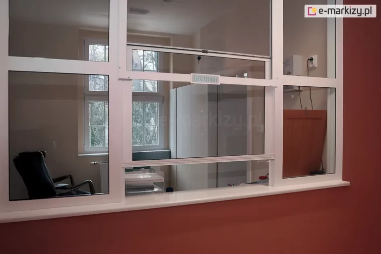 Biała zabudowa okienna typu recepcja, wykonaną z profili aluminiowych z przeszkleniem. Centralnym elementem jest poziome, przesuwne okienko podawcze z napisem "RECEPTION" umieszczonym na ramie, zabezpieczone małym zamkiem punktowym w górnej części. Konstrukcja osadzona jest na ścianie o koralowym lub ceglastym kolorze, wykończonej białym parapetem. Przez szyby widać wnętrze biura z ciemnym fotelem biurowym, szafami dokumentowymi oraz klasycznym, białym oknem w tle, przez które prześwitują drzewa.