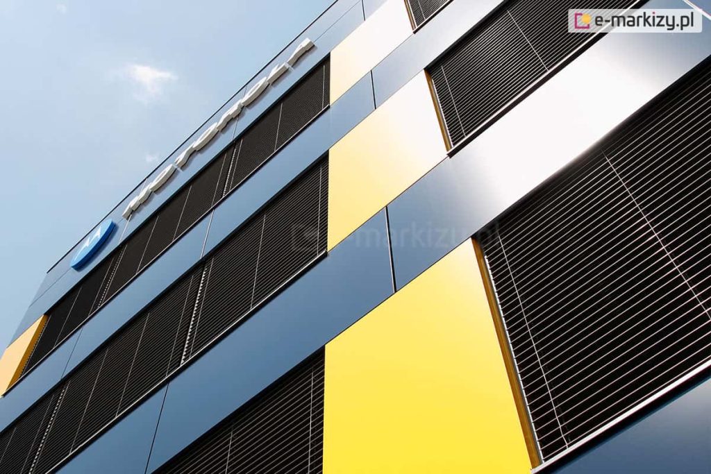 Perspektywiczne ujęcie narożnika nowoczesnego budynku biurowego z fasadą wentylowaną. Elewacja składa się z dużych paneli w kolorze granatowym (grafitowym) oraz kontrastujących, pionowych pasów w kolorze intensywnie żółtym. Na całej wysokości przeszkleń zainstalowano zewnętrzne żaluzje fasadowe w kolorze czarnym lub bardzo ciemnym graficie. Lamele są ustawione pod kątem, chroniąc wnętrze przed słońcem, jednocześnie odbijając światło dzienne. Na górnej części elewacji widoczny jest fragment logotypu korporacyjnego montowanego przestrzennie. Zdjęcie wykonane pod kątem ku górze na tle bezchmurnego, jasnego nieba.