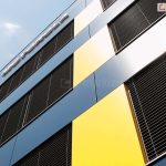 Perspektywiczne ujęcie narożnika nowoczesnego budynku biurowego z fasadą wentylowaną. Elewacja składa się z dużych paneli w kolorze granatowym (grafitowym) oraz kontrastujących, pionowych pasów w kolorze intensywnie żółtym. Na całej wysokości przeszkleń zainstalowano zewnętrzne żaluzje fasadowe w kolorze czarnym lub bardzo ciemnym graficie. Lamele są ustawione pod kątem, chroniąc wnętrze przed słońcem, jednocześnie odbijając światło dzienne. Na górnej części elewacji widoczny jest fragment logotypu korporacyjnego montowanego przestrzennie. Zdjęcie wykonane pod kątem ku górze na tle bezchmurnego, jasnego nieba.