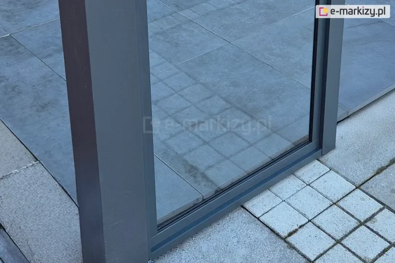 Detal dolnej części okna stałego systemu Fest, dedykowanego do zabudowy tarasów i zadaszeń. Widoczny jest solidny, pionowy profil aluminiowy oraz dolna rama w kolorze antracytowym, utrzymująca taflę szkła. Konstrukcja jest zamontowana bezpośrednio przy nawierzchni wykonanej z dużych, szarych płyt tarasowych, które sąsiadują z mniejszą kostką brukową w jasnym odcieniu.