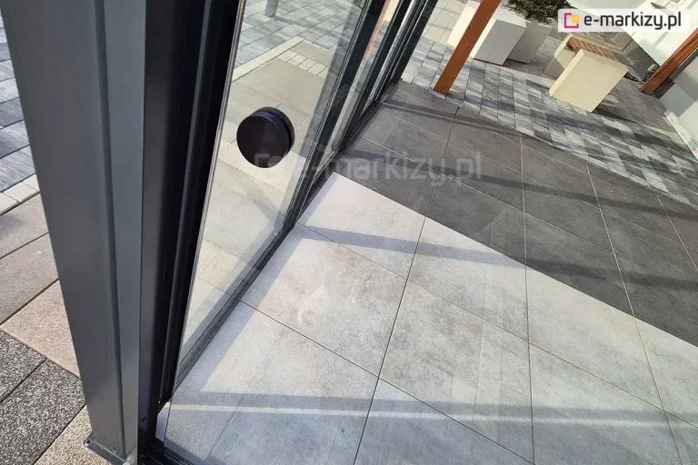 Szklany panel przesuwny bezramowy Miros zamocowany do zadaszenia tarasowego Modes na podłożu z płyt betonowych wyposażony został w czarny okrągły uchwyt ułatwiający przesuwanie panelu po torze.
