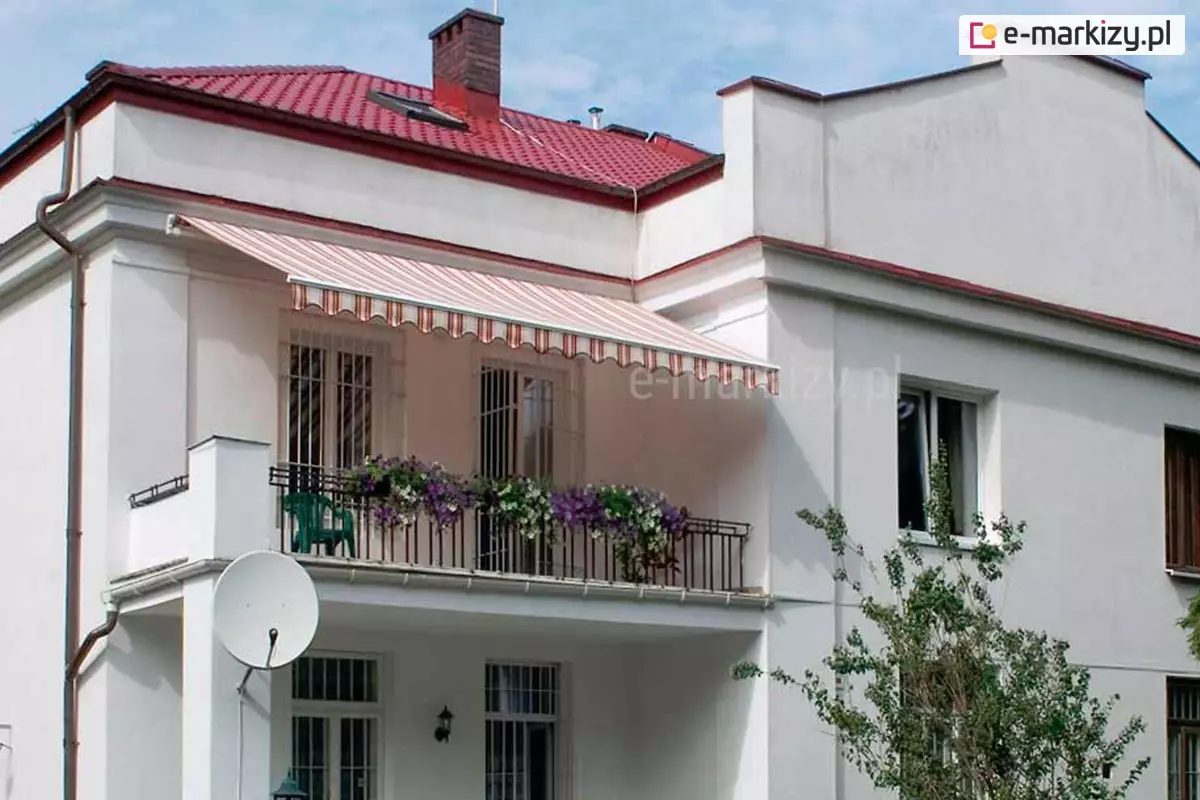 Zdjęcie przedstawia szerokie ujęcie elewacji białego, piętrowego budynku mieszkalnego z dachem krytym czerwoną blachodachówką, na którym zainstalowana została markiza bez kasety Jamaica. Osłona przeciwsłoneczna jest w pełni rozwinięta, osłaniając przestronny balkon na piętrze, zabezpieczony czarną, metalową balustradą ozdobioną donicami z fioletowymi i białymi kwiatami. Poszycie markizy wykonane jest z wysokiej jakości tkaniny w klasyczne pionowe pasy w odcieniach bieli, beżu i czerwieni, co estetycznie nawiązuje do koloru dachu budynku. Frontowa część poszycia wykończona jest ozdobną falbaną o falistym kształcie z dopasowaną kolorystycznie lamówką. Konstrukcja wsporcza, którą posiada markiza bez kasety Jamajka, jest biała i wtapia się w kolorystykę jasnej ściany, a jej ramiona przegubowe są niewidoczne pod materiałem, co nadaje całości lekkiego wyglądu. Montaż został wykonany powyżej wnęki balkonowej, zapewniając skuteczną ochronę przed słońcem dla drzwi balkonowych oraz wnętrza pomieszczenia. W dolnej części kadru widoczny jest fragment parteru z białą anteną satelitarną zamocowaną do filaru podtrzymującego płytę balkonu oraz nasadzenia ogrodowe w postaci drzew i krzewów.