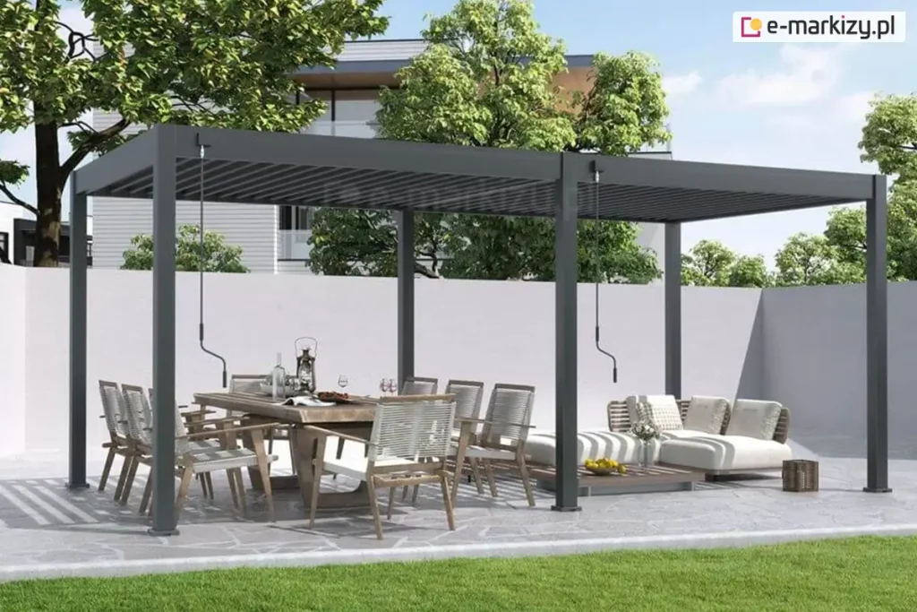 Wolnostojąca pergola tarasowa Marlo o konstrukcji dwumodułowej w kolorze ciemnoszarym (antracytowym), zamontowana na tarasie wyłożonym nieregularnymi płytami kamiennymi. Całość opiera się na sześciu aluminiowych słupach o kwadratowym przekroju, a zadaszenie stanowią ruchome lamele, które w lewej sekcji są lekko uchylone, natomiast w prawej pozostają w pozycji otwartej, przepuszczając naturalne światło. Przy słupach konstrukcyjnych widoczne są dwa oddzielne mechanizmy korbowe służące do manualnego sterowania każdą z sekcji dachu niezależnie. Pod lewym modułem ustawiono duży stół jadalniany z masywnym blatem oraz krzesła o białym splocie z szarymi poduszkami. Pod prawym modułem znajduje się strefa relaksu z niską, białą sofą modułową i dopasowanym stolikiem kawowym. W tle widoczny jest wysoki, jasny mur odgradzający posesję, za którym rosną gęste korony drzew liściastych, a w oddali zarysowuje się bryła nowoczesnego budynku. Przed tarasem rozciąga się pas równo przystrzyżonego, zielonego trawnika.