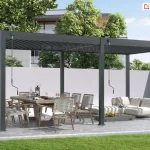 Wolnostojąca pergola tarasowa Marlo o konstrukcji dwumodułowej w kolorze ciemnoszarym (antracytowym), zamontowana na tarasie wyłożonym nieregularnymi płytami kamiennymi. Całość opiera się na sześciu aluminiowych słupach o kwadratowym przekroju, a zadaszenie stanowią ruchome lamele, które w lewej sekcji są lekko uchylone, natomiast w prawej pozostają w pozycji otwartej, przepuszczając naturalne światło. Przy słupach konstrukcyjnych widoczne są dwa oddzielne mechanizmy korbowe służące do manualnego sterowania każdą z sekcji dachu niezależnie. Pod lewym modułem ustawiono duży stół jadalniany z masywnym blatem oraz krzesła o białym splocie z szarymi poduszkami. Pod prawym modułem znajduje się strefa relaksu z niską, białą sofą modułową i dopasowanym stolikiem kawowym. W tle widoczny jest wysoki, jasny mur odgradzający posesję, za którym rosną gęste korony drzew liściastych, a w oddali zarysowuje się bryła nowoczesnego budynku. Przed tarasem rozciąga się pas równo przystrzyżonego, zielonego trawnika.