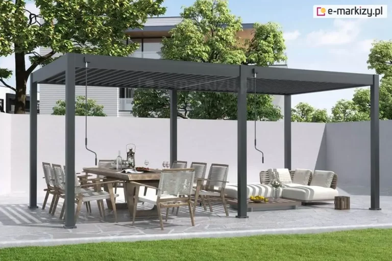Wolnostojąca pergola tarasowa Marlo o konstrukcji dwumodułowej w kolorze ciemnoszarym (antracytowym), zamontowana na tarasie wyłożonym nieregularnymi płytami kamiennymi. Całość opiera się na sześciu aluminiowych słupach o kwadratowym przekroju, a zadaszenie stanowią ruchome lamele, które w lewej sekcji są lekko uchylone, natomiast w prawej pozostają w pozycji otwartej, przepuszczając naturalne światło. Przy słupach konstrukcyjnych widoczne są dwa oddzielne mechanizmy korbowe służące do manualnego sterowania każdą z sekcji dachu niezależnie. Pod lewym modułem ustawiono duży stół jadalniany z masywnym blatem oraz krzesła o białym splocie z szarymi poduszkami. Pod prawym modułem znajduje się strefa relaksu z niską, białą sofą modułową i dopasowanym stolikiem kawowym. W tle widoczny jest wysoki, jasny mur odgradzający posesję, za którym rosną gęste korony drzew liściastych, a w oddali zarysowuje się bryła nowoczesnego budynku. Przed tarasem rozciąga się pas równo przystrzyżonego, zielonego trawnika.
