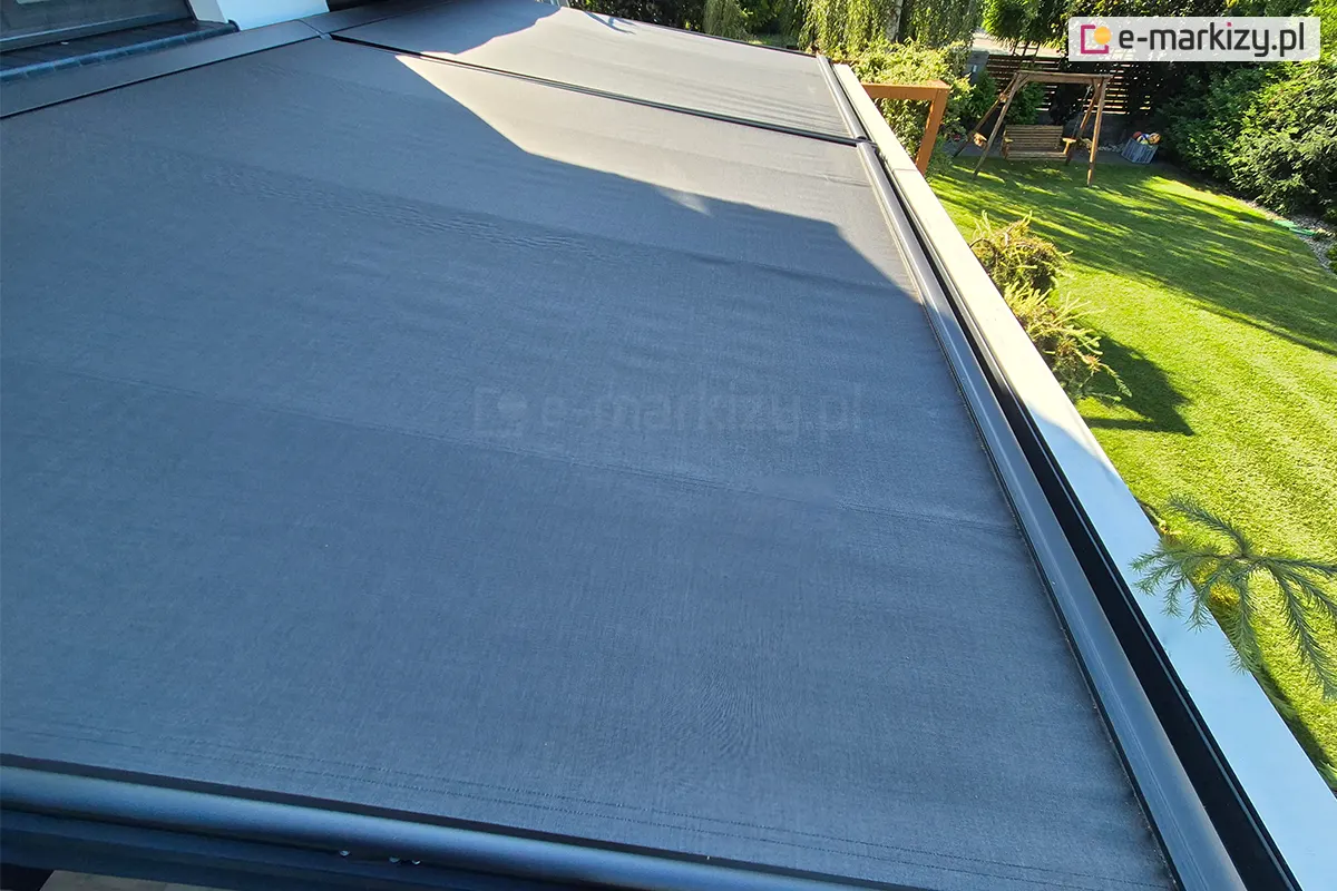 Widok z góry na rozłożoną, nowoczesną roletę veranda w kolorze ciemnoszarym lub antracytowym, która została zamontowana nad przydomowym tarasem. Poszycie jest wykonane z gęsto tkanego materiału o gładkiej strukturze, który wypełnia przestrzeń między prowadnicami bocznymi. Tkanina jest napięta i podzielona na dwie niezależne sekcje przez poprzeczne prowadnice, co zapewnia stabilność konstrukcji. Po prawej stronie widoczna jest metalowa belka dolna oraz fragment rynny lub obróbki blacharskiej wykończonej w kolorze dopasowanym do poszycia. Cień rzucany przez dach na zadaszenie pokrywa niemal całą powierzchnię materiału, podkreślając jego funkcję ochronną przed słońcem. W tle po prawej stronie rozciąga się zadbany ogród z soczyście zielonym trawnikiem, na którym znajduje się drewniana huśtawka ogrodowa oraz różnorodne krzewy i drzewa iglaste. Światło słoneczne oświetla krawędź ogrodu, tworząc wyraźny kontrast między zacienioną strefą wypoczynku pod verandą a nasłonecznioną częścią rekreacyjną posesji.