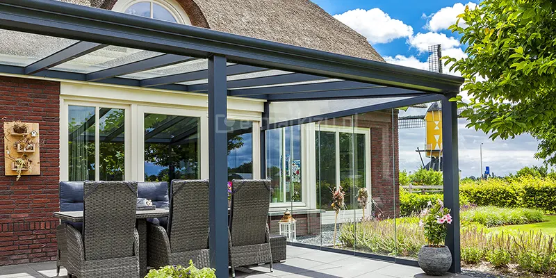 Zdjęcie prezentuje narożną sekcję zadaszenia tarasowego model Terano, zamontowaną przy tradycyjnym budynku z czerwonej cegły, krytym strzechą. Konstrukcja zadaszenia wykonana jest z profili aluminiowych w kolorze antracytowym. Front dachu wieńczy profil rynnowy o zaokrąglonym kształcie, wsparty na pionowym słupie nośnym. Ściana boczna zadaszenia została w pełni zabudowana systemem przesuwnych paneli szklanych, które tworzą transparentną osłonę przed wiatrem. Ponad przesuwnymi drzwiami, w trójkątnym polu pod skosem dachu, umieszczono stałe wypełnienie szklane, tzw. klin. Pod zadaszeniem znajduje się strefa jadalniana z meblami z technorattanu w kolorze szarym – widoczne są wysokie oparcia krzeseł oraz fragment stołu. W tle, za szklaną zabudową, rozpościera się widok na ogród z lawendą oraz charakterystyczny żółto-czarny wiatrak. Całość kompozycji dopełniają donice z kwiatami oraz jasna, płytowa nawierzchnia tarasu.