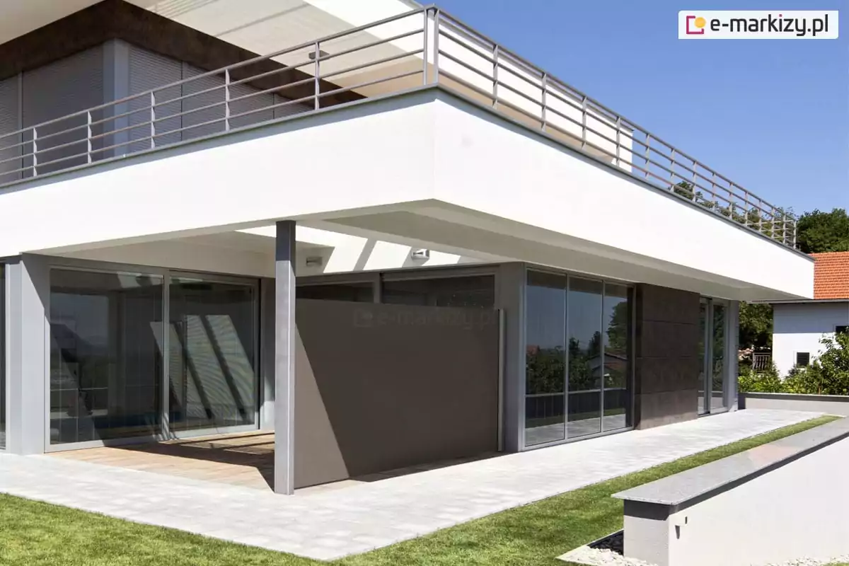 Nowoczesna, wielopoziomowa bryła budynku mieszkalnego o minimalistycznym charakterze, zdominowana przez biel elewacji i duże przeszklenia. Na parterze znajduje się przestronny, zadaszony taras, którego sufit stanowi jednocześnie konstrukcję balkonu górnej kondygnacji. W centralnej części tarasu, pomiędzy stalowym filarem konstrukcyjnym a ścianą budynku, zamontowano boczną markizę Bora wyciąganą w kolorze szarobrązowym lub taupe. Markiza jest w pełni rozwinięta, tworząc jednolitą, nieprzezierną płaszczyznę, która dzieli przestrzeń tarasu i zapewnia osłonę przed wzrokiem oraz wiatrem. Posadzka tarasu jest dwudzielna – część pod zadaszeniem wyłożona jest drewnianymi deskami o naturalnym odcieniu, natomiast zewnętrzna strefa komunikacyjna oraz opaska wokół domu wykonane są z jasnoszarej kostki brukowej lub płyt betonowych. Budynek posiada liczne, wysokie okna o szarych profilach, które odbijają otoczenie. Na piętrze widoczny jest długi taras zabezpieczony prostą, stalową balustradą składającą się z poziomych prętów. Architektura domu wykorzystuje również ciemne akcenty w postaci grafitowych płyt elewacyjnych na fragmencie parteru. Przed budynkiem rozciąga się równo przystrzyżony, zielony trawnik, a wzdłuż tarasu przebiega wąski pas niskiej roślinności lub dekoracyjnego żwiru. Scena osadzona jest w słoneczny dzień, przy bezchmurnym, błękitnym niebie, co podkreśla czystość formy architektonicznej.