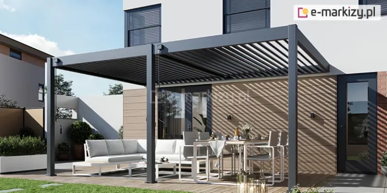 Nowoczesna pergola tarasowa Rubio Soft, o konstrukcji aluminiowej w kolorze antracytowym zamontowana przy elewacji piętrowego budynku mieszkalnego. Zadaszenie pergoli składa się z ruchomych lameli, które są ustawione pod kątem, co pozwala na regulację natężenia światła słonecznego padającego na taras. Konstrukcja wsparta jest na solidnych, kwadratowych słupach, a przy dwóch z nich widoczne są mechanizmy korbowe służące do ręcznego sterowania dachem. Pod pergolą zaaranżowana została strefa wypoczynkowa oraz jadalniana na drewnianym podeście. Po lewej stronie znajduje się biały narożnik ogrodowy z jasnoszarymi poduszkami oraz niski stolik kawowy. Po prawej stronie ustawiono duży stół obiadowy z białym blatem, przy którym stoją nowoczesne krzesła z szarymi siedziskami. Na stole rozłożona jest zastawa, szklanki oraz dekoracje w postaci wazonu z kwiatami. Elewacja domu w tle łączy biały tynk z panelami o strukturze drewna w jasnym odcieniu. Cień rzucany przez lamele tworzy na ścianie budynku regularny wzór ukośnych pasów. W otoczeniu tarasu widoczna jest starannie wypielęgnowana zieleń w białych donicach oraz fragment trawnika. Całość przedstawiona jest w pełnym świetle dziennym przy bezchmurnym niebie.