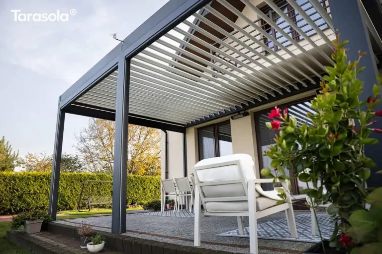 Zdjęcie przedstawia nowoczesną pergolę Essential Tarasola w kolorze ciemnoantracytowym, uwiecznioną z niskiej perspektywy, co eksponuje zaawansowaną konstrukcję dachu lamelowego. Aluminiowe lamele w kolorze białym są częściowo uchylone, ukazując precyzję mechanizmu obrotowego, który pozwala na kontrolowanie dopływu światła słonecznego do wnętrza tarasu. Rama zadaszenia Essential Tarasola spoczywa na solidnych, pionowych słupach o kwadratowym przekroju, a na górnej belce widoczny jest niewielki czujnik pogodowy. Pod pergolą znajduje się komfortowa strefa wypoczynkowa; na pierwszym planie widoczny jest tył białego fotela ogrodowego z miękką poduszką, ustawionego na dywanie zewnętrznym w szaro-niebieskie geometryczne wzory. W głębi, pod jasną elewacją domu, znajduje się zestaw jadalniany z białym stołem i nowoczesnymi krzesłami. Na ścianie budynku, tuż nad linią wzroku, zamontowany jest podłużny promiennik ciepła. Po prawej stronie kadru, na bardzo bliskim planie, widoczna jest zielona roślina doniczkowa z wyraźnymi, czerwonymi kwiatami, która dodaje kompozycji naturalnego akcentu. W tle rozciąga się zadbany ogród z zielonym żywopłotem, huśtawką oraz drzewami o jesiennym zabarwieniu liści. Jasna elewacja budynku z brązowymi ramami okiennymi oraz fragment balkonu z ciemną balustradą dopełniają estetykę całej realizacji Essential Tarasola.